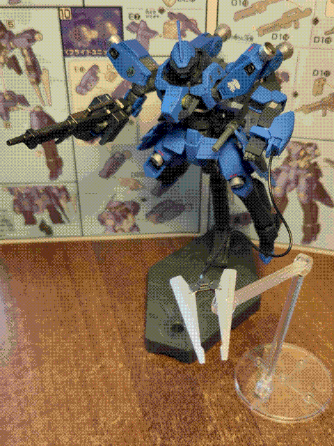 A photo of the high-grade McGillis' Schwalbe Graze model kit, posed in the air on a stand. It is a blue mech with sharp, angular knight-like armor panels and a skeletal frame. The head has two narrow slits for optical sensors, one above the other, and it has thrusters embedded in the knees, shoulders, and on 'arms' on the back of the mobile suit. It has a decal of the Gjallarhorn crest on its left shoulder, and the Fareed family crest on its right. It is holding a rifle, has an axe at its side, and is posed to be 'shooting' its wire blade out (which is perched on another stand to hold it in the air). The instruction manual for the kit is visible in the background.