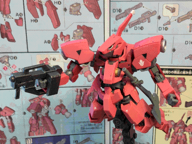 A photo of the high-grade Ryusei-Go (Graze Custom II) model kit, posed in the air on a stand. It is a red-pink mech with sharp, knight-like armor panels (except for the shoulders, which come from a different mech in the fiction of the show) and a skeletal frame. The head has a rectangular aperture for a singular optical sensor, and the left shoulder has a white fleur-de-lis-like symbol on it (the Tekkadan symbol). There are also eyes 'painted' (actually an applied decal) on either side of the head. It is holding a short sawn-off rifle in its right hand, while its left hand is empty (although it has an axe at its side). The instruction manual for the kit is visible in the background.