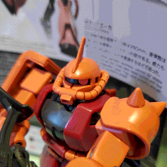 A photo of the Char's Zaku II model kit, zoomed in on the head. Using a combination of the head itself rotating and the monoeye moving in its track, the monoeye is pointed directly at the camera. The head resembles a gas mask, with a long 'snout' intake connected to coolant tubes that run around the head. It also has an antenna on the top, reflecting it being a commander model.