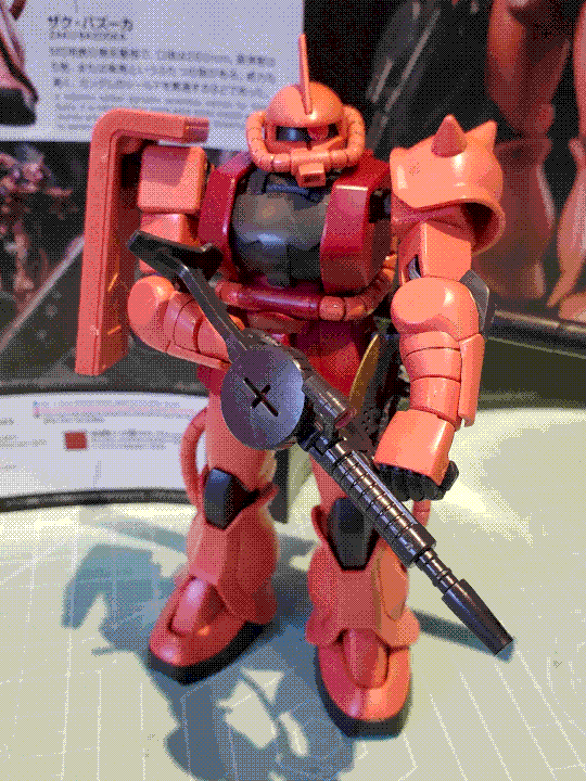 A photo of the Char's Zaku II high-grade model kit ('revive' revision). It is a bulky humanoid mech, with curved red armor pieces and coolant tubes around the waist, legs, and helmet-like head. It has a single pink monoeye that can move left to right in a track, a spiked armor plate on its left shoulder, and a large shoulder shield on its right shoulder. It is holding a machine gun in both hands, and the instruction manual is visible in the background.