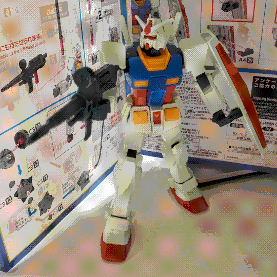 A photo of the RX-78-2 Gundam entry-grade model kit. It is similar in appearance to the Beyond Global high-grade kit, being a humanoid mech that is mostly white with red, blue, and yellow details. However, the white is a much cooler color temperature and the other colours are more saturated. It is holding a beam rifle in its right hand and a shield in its right, and is posed as if shooting at something to the left of the photo frame. The instruction manual is visible in the background.