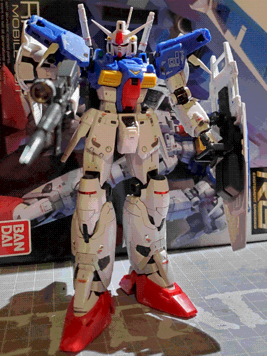 A photo of the real-grade GP01-Fb model kit. It is a humanoid mech with a white lower body, blue upper body, and red and yellow accents. It has somewhat bulky, bulbous armor, and has vernier thrusters in several locations including two massive boosters on its back. It is posed looking down at the camera, with its rifle pointed in the same direction. The box art is visible in the background.