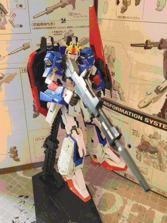 A photo of the real-grade Zeta Gundam model kit. It is a humanoid mech with white, red, white, and yellow panels. It also has a large 'backpack' and a thin shield on its left arm, both red and black. It has thruster vents everywhere, most notably on the shoulders and ankles, and is holding a large beam rifle in its right hand. It is posed on a raised stand, leaned slightly backwards and head looking towards the camera. The instruction manual for the kit is in the background.