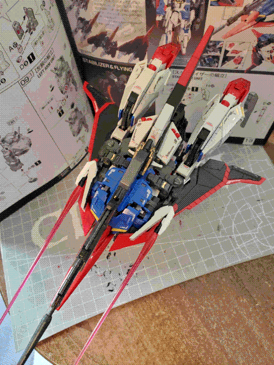 A photo of the Zeta Gundam real-grade model kit in its 'Waverider' mode. The chest has been flipped out and its legs have been brought inwards to form the rear thrusters, while the backpack and shield have been attached underneath to provide an aerodynamic wing surface. The beam rifle is attached in the centre, on top of the flipped-out chest piece. The beam sabers in the side-skirt armor have also been exposed so they can act as side-mounted beam rifles. The photo is taken from a mostly overhead view, and the instruction manual is visible in the background.