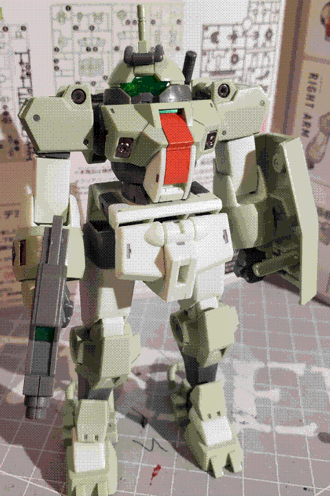 A photo of the Demi Trainer high-grade model kit. It is a squat, humanoid mech similar in appearance to Chuchu's Demi Trainer, although it has a more uniform appearance overall and a white-and-light-green color palette, giving it a cooler appearance in terms of colour temperature. The domed head has wide optical panel, and the cockpit hatch is colored bright red. It is holding a short, boxy beam rifle in its right hand and a chunky shield in its left hand.