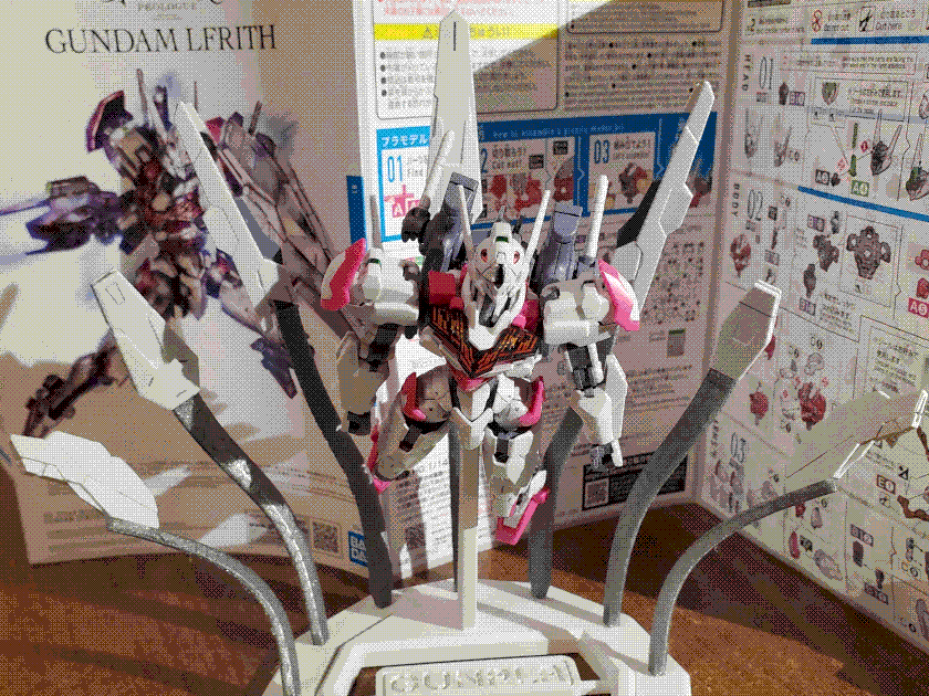 A picture of a plastic model kit of the mech Gundam Lfrith from Mobile Suit Gundam: The Witch From Mercury. The mech is arranged on a stand to appear as if in flight: behind it are the 'GUND Bits' that make up its shield spread outwards in a fan. The picture is head-on. The assembly manual for the kit is behind it, taking up the entire background.